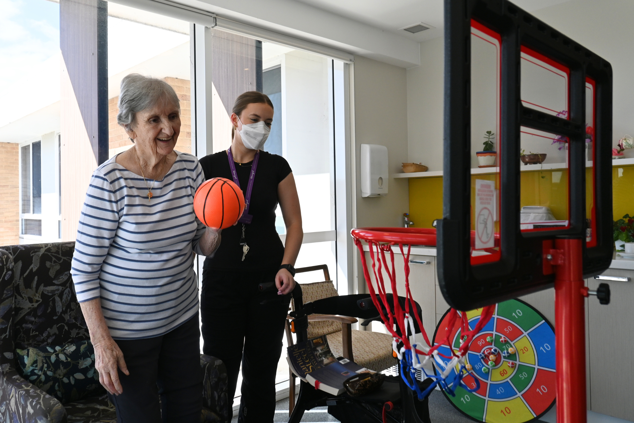 Warrigal aged care resident at Shell Cove in a striped top is holding a basketball and smiling with a staff member nearby